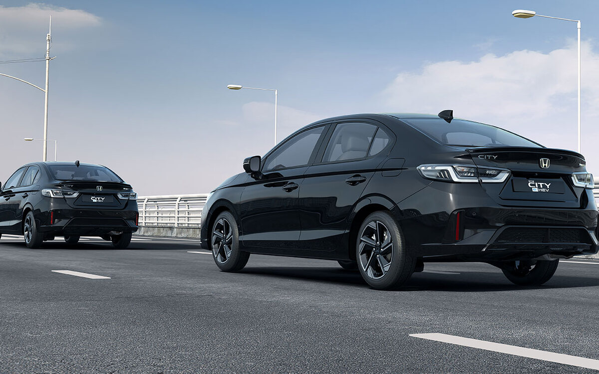 Two black facelifted Honda City e:HEV sedans on a bridge road under a blue sky, seen from the rear three-quarter, one following the other past a traffic light