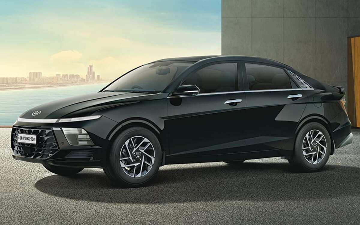 A black Hyundai Verna parked on an open forecourt beside a concrete and wood-panelled building, seen from the front-left side, with a waterfront city skyline behind it