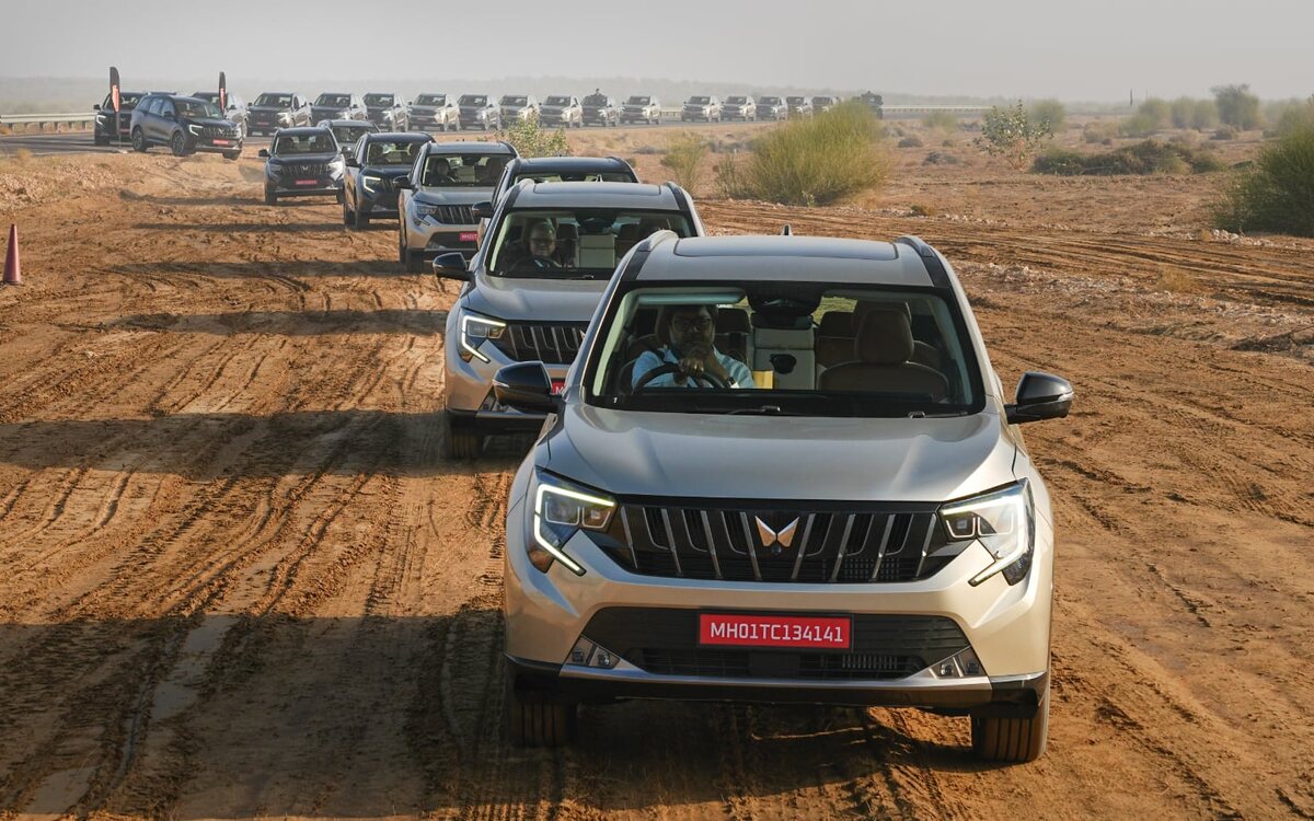 A silver Mahindra XUV 7XO seen head-on leading a convoy of XUV 7XOs along a rutted sandy desert track, with a line of more 7XOs on the road behind