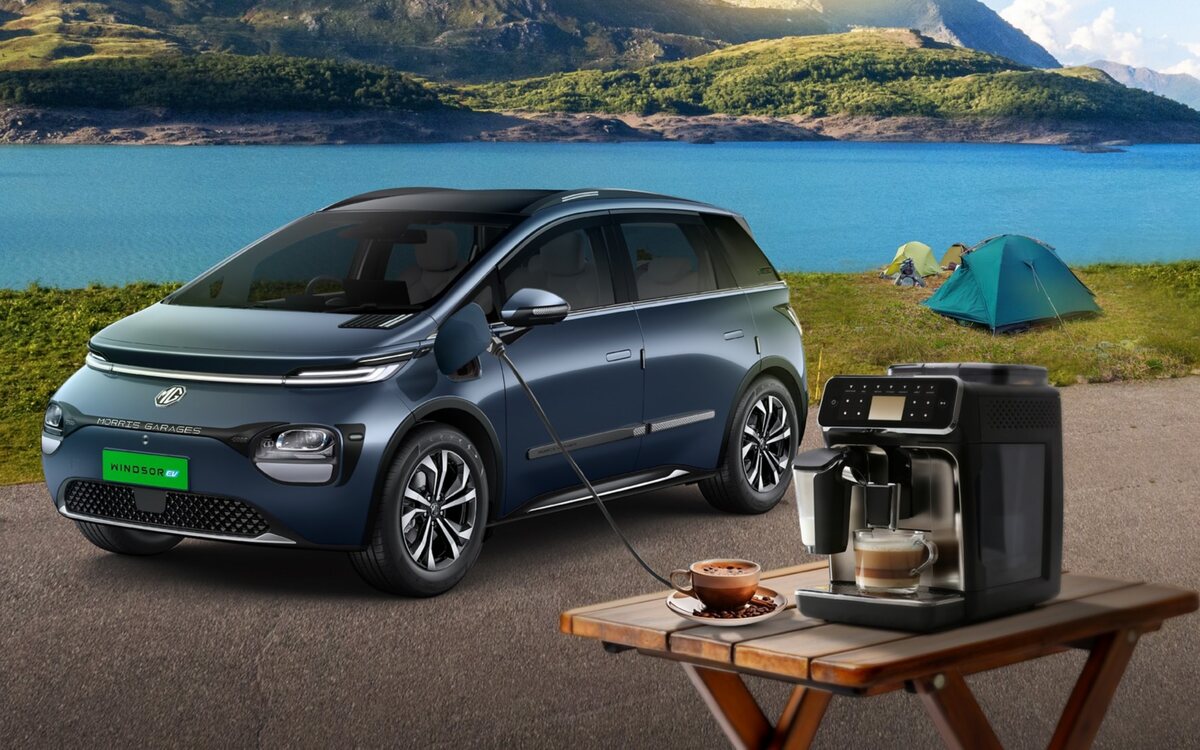 A dark blue MG Windsor EV parked on a lakeside road with green hills behind, a cable from its charging port powering a coffee machine on a folding wooden table, with tents pitched on the grass
