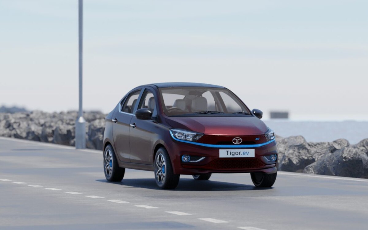A Magnetic Red Tata Tigor EV with its blue front light strip and Tigor.ev plate, driving on a seaside road past a rock embankment and a lamp post under a hazy sky