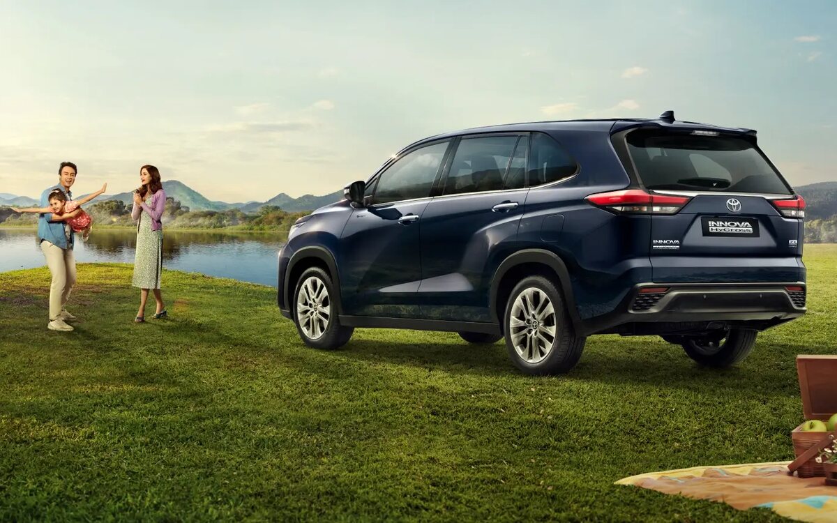 A dark blue Toyota Innova HyCross parked on a grassy lakeside field, seen from the rear three-quarter, with a family playing beside it