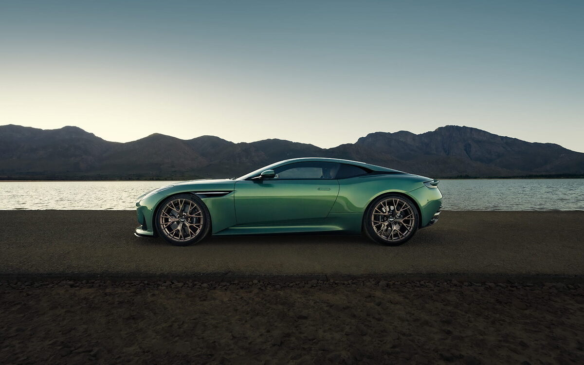 Green Aston Martin DB12 in side profile beside a lake with mountains behind
