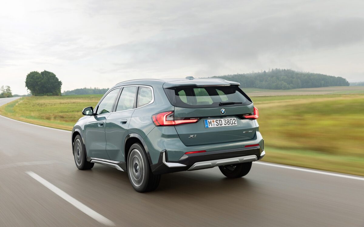 BMW X1 sDrive18d in teal-grey paint seen from the rear three-quarter while driving along a road