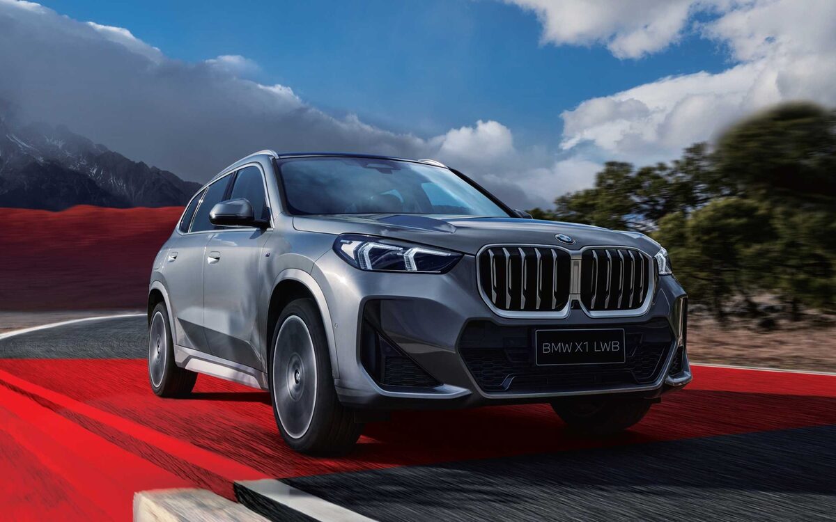 BMW X1 LWB in grey, front three-quarter view driving on a red-edged track with mountains behind