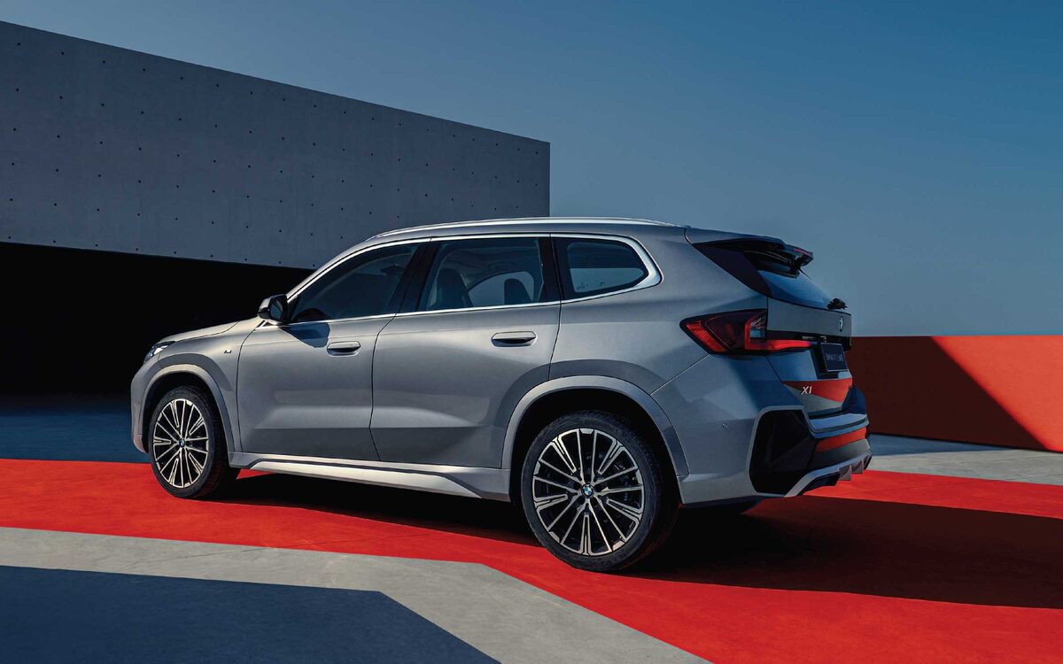BMW X1 LWB in light grey, side and rear three-quarter view beside a concrete building on a red and grey forecourt