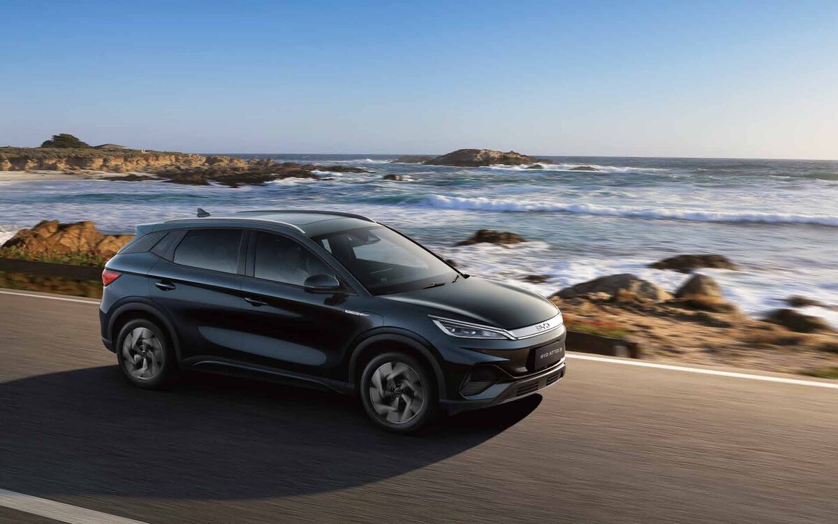 BYD Atto 3 in black driving along a coastal road with rocks and sea behind