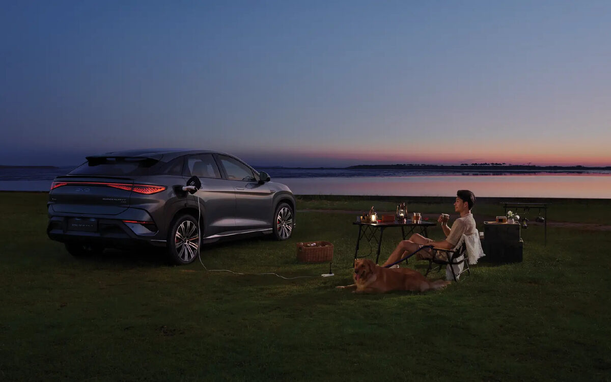 BYD Sealion 7 parked on a lakeside lawn at dusk, charging a camp table with its vehicle-to-load feature