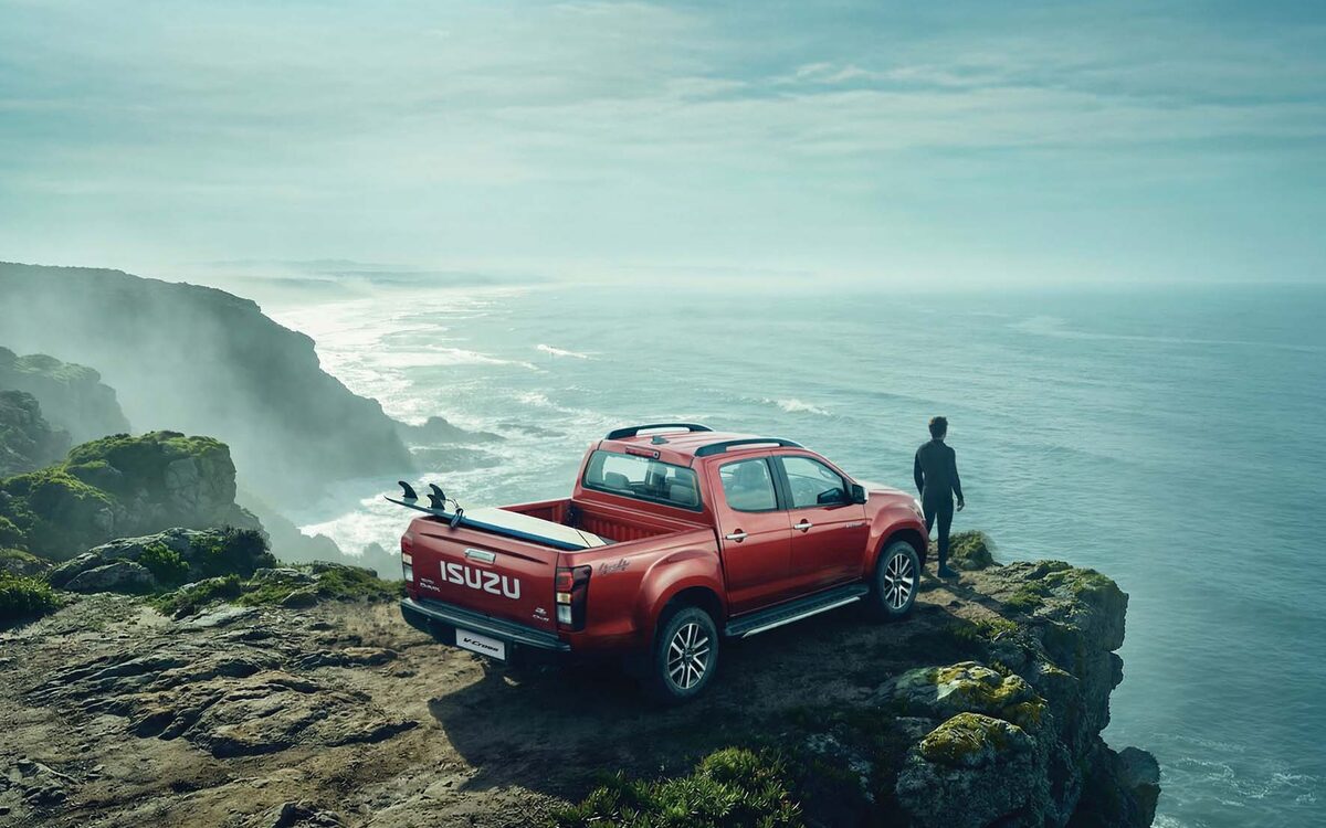 Isuzu D-MAX V-Cross in red parked on a coastal cliff with a person looking at the sea, rear three-quarter view