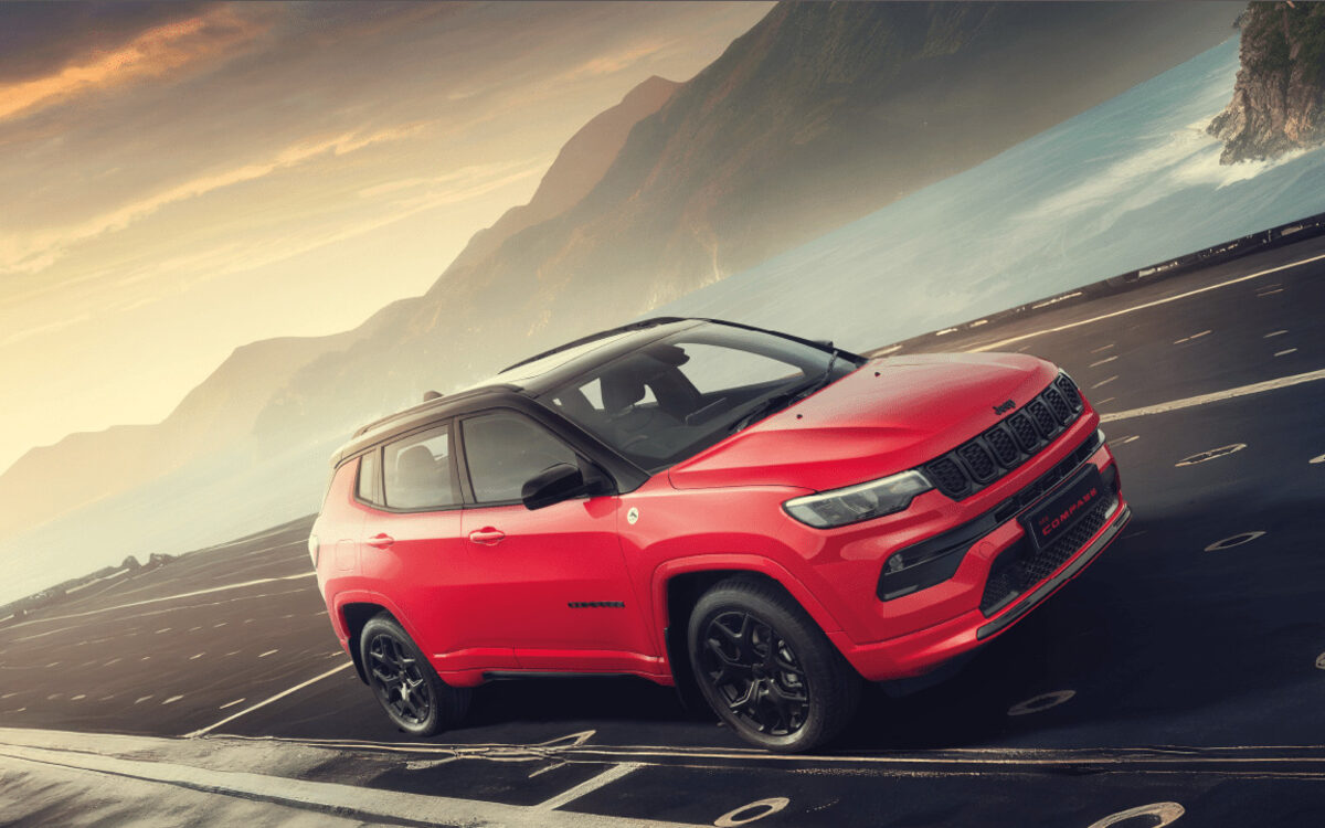 Red Jeep Compass with black roof driving on a mountain road at sunset