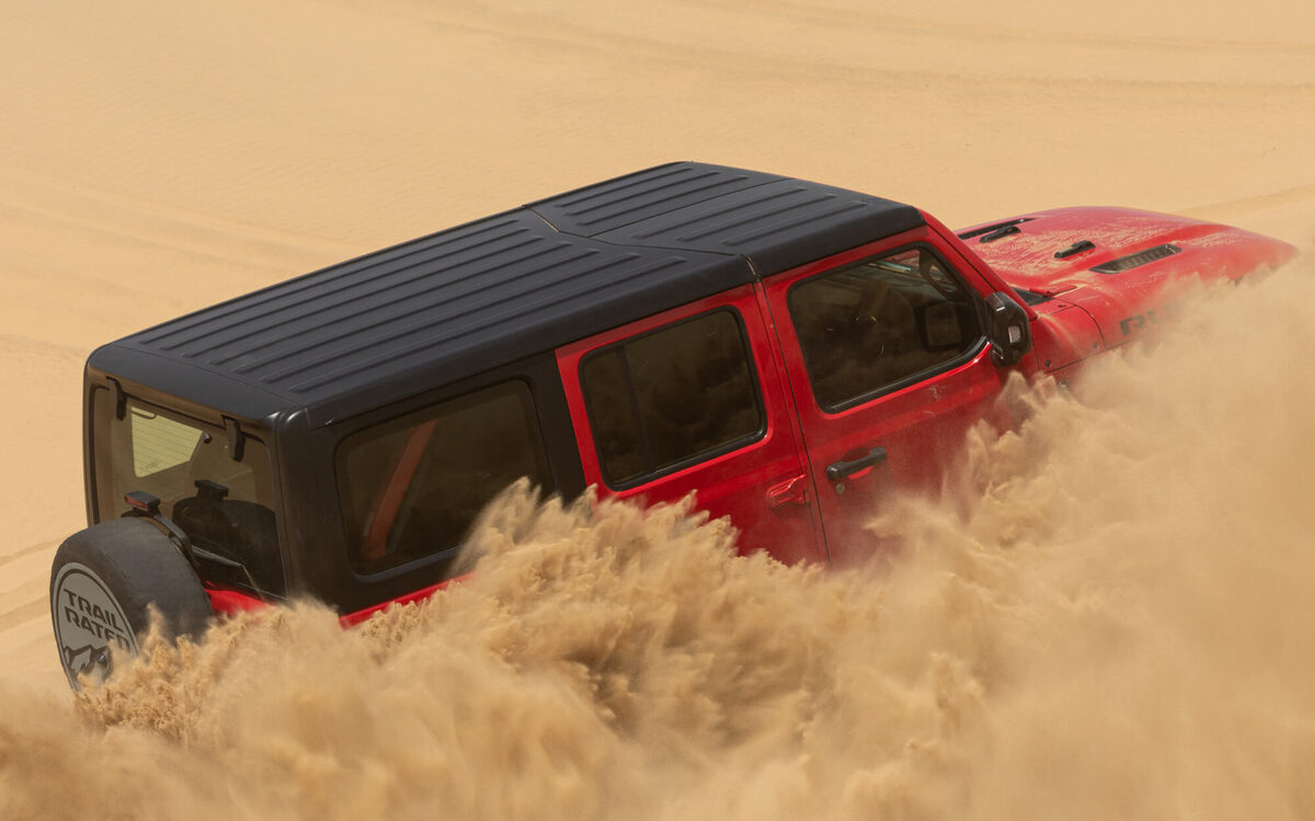 Jeep Wrangler in red with a black hard top, rear three-quarter view driving through sand