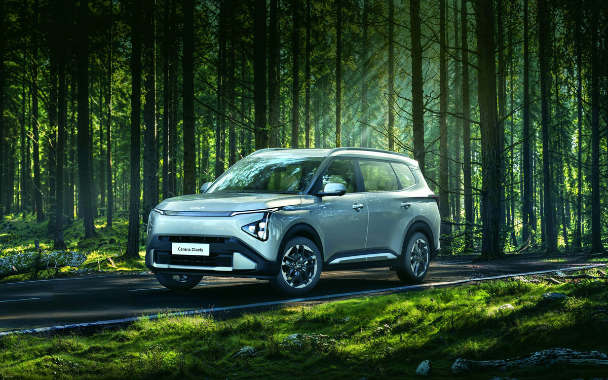 Kia Carens Clavis in a pale grey-green shade driving along a forest road, front three-quarter view