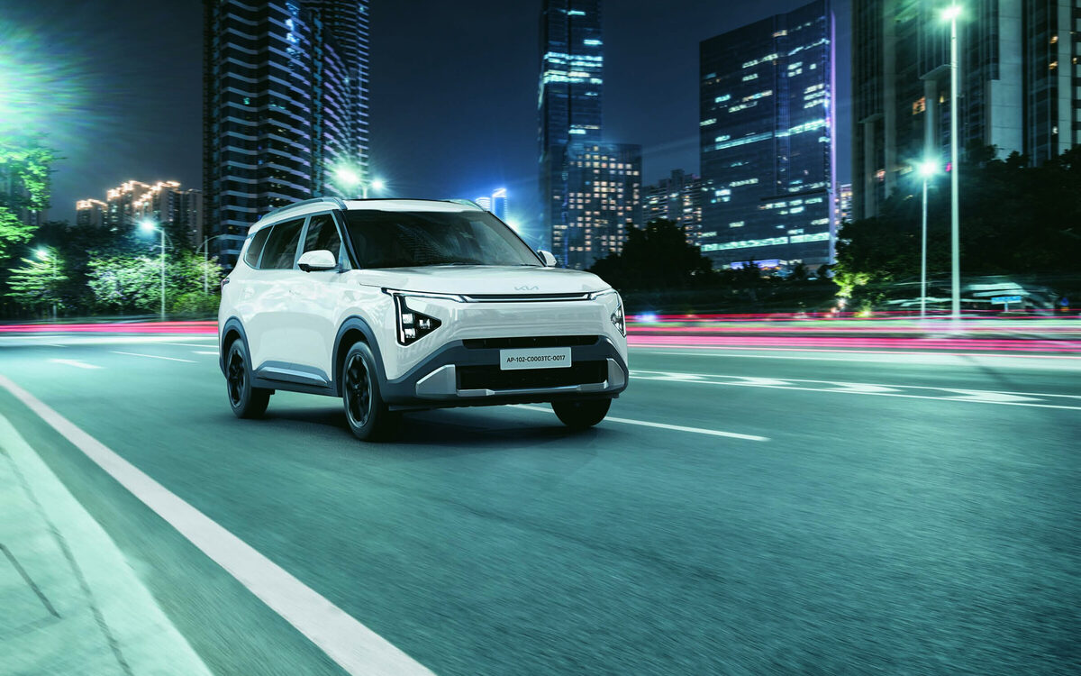 White Kia Carens Clavis driving along a city road at night with tall buildings behind
