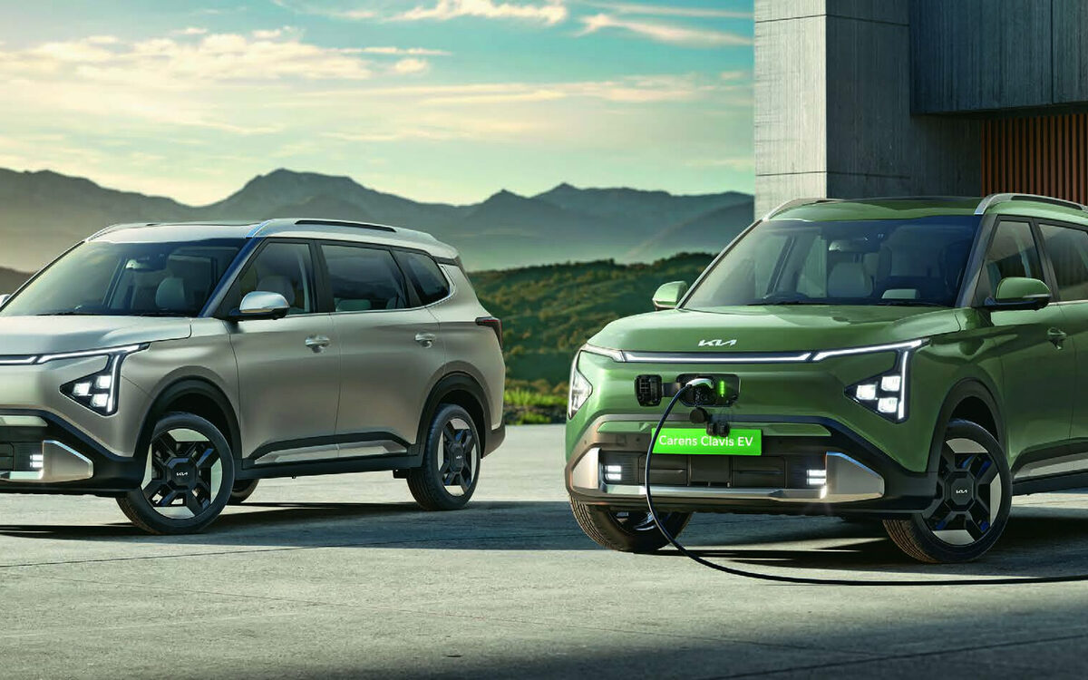 Two Kia Carens Clavis EVs parked in front of a house, the green one plugged into a wall charger