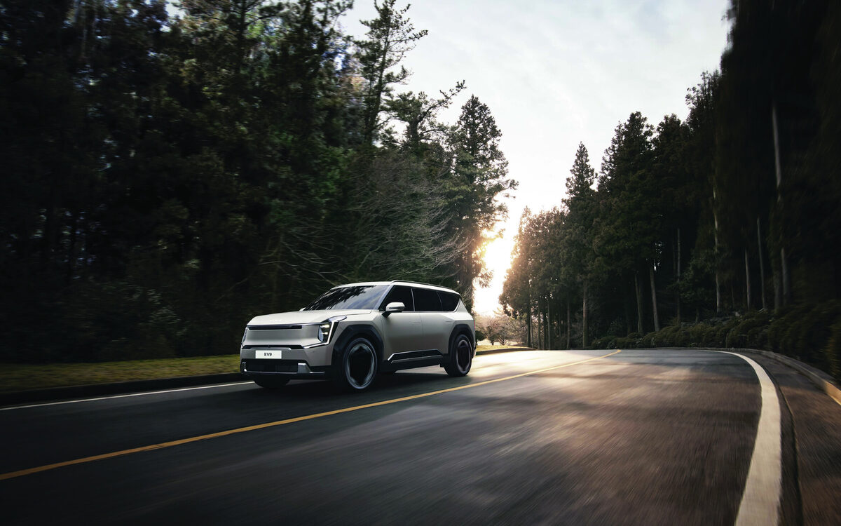 Kia EV9 in light grey driving along a forest road in low sun