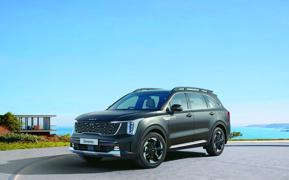 Kia Sorento Hybrid in dark grey, front three-quarter view on a curving seaside road with a glass house and sea behind