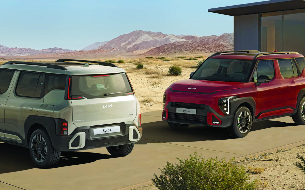 Two Kia Syros SUVs, one in light green and one in red, parked on a desert road beside a glass house