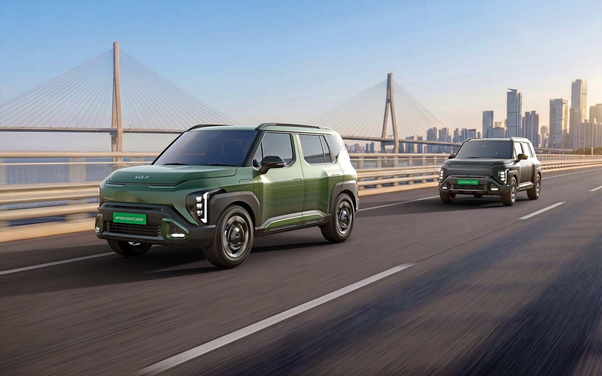Two Kia Syros EV cars, a green one in front, driving on a road near a cable-stayed bridge