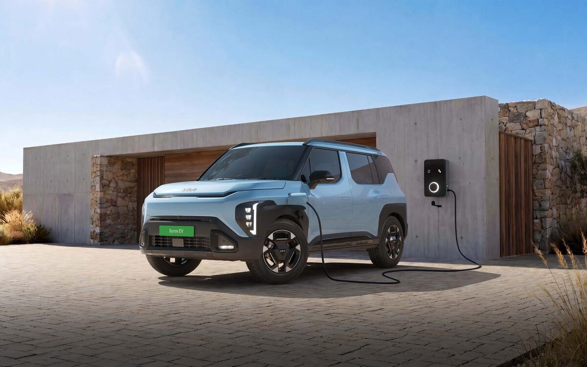Kia Syros EV in light blue plugged into a wall charger outside a stone and concrete house