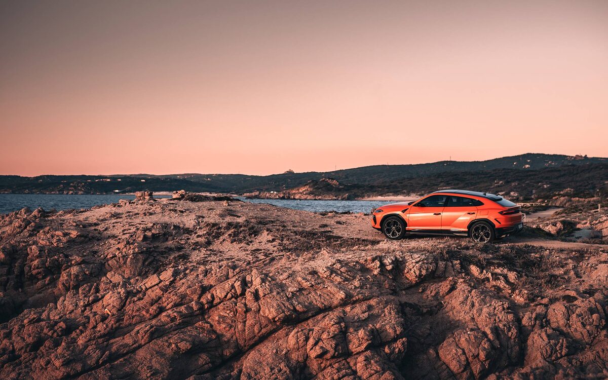 Lamborghini Urus SE in orange parked on rocky ground by the sea at sunset