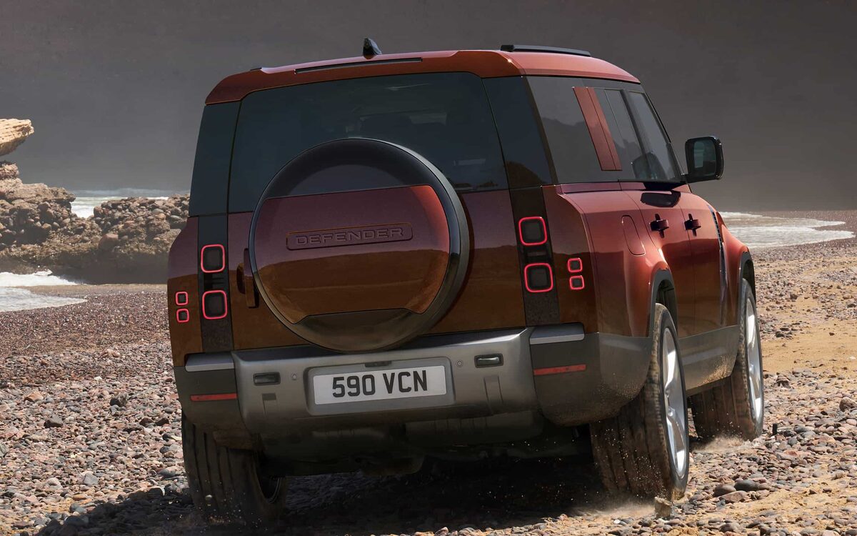 Land Rover Defender in red with spare wheel, rear three-quarter view on a gravel beach