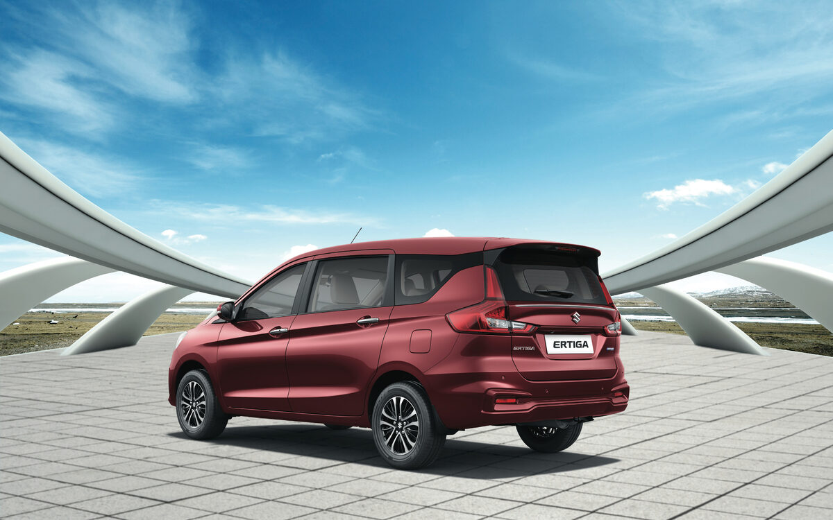 Maruti Suzuki Ertiga in Auburn Red, rear three-quarter view on a paved plaza under a blue sky