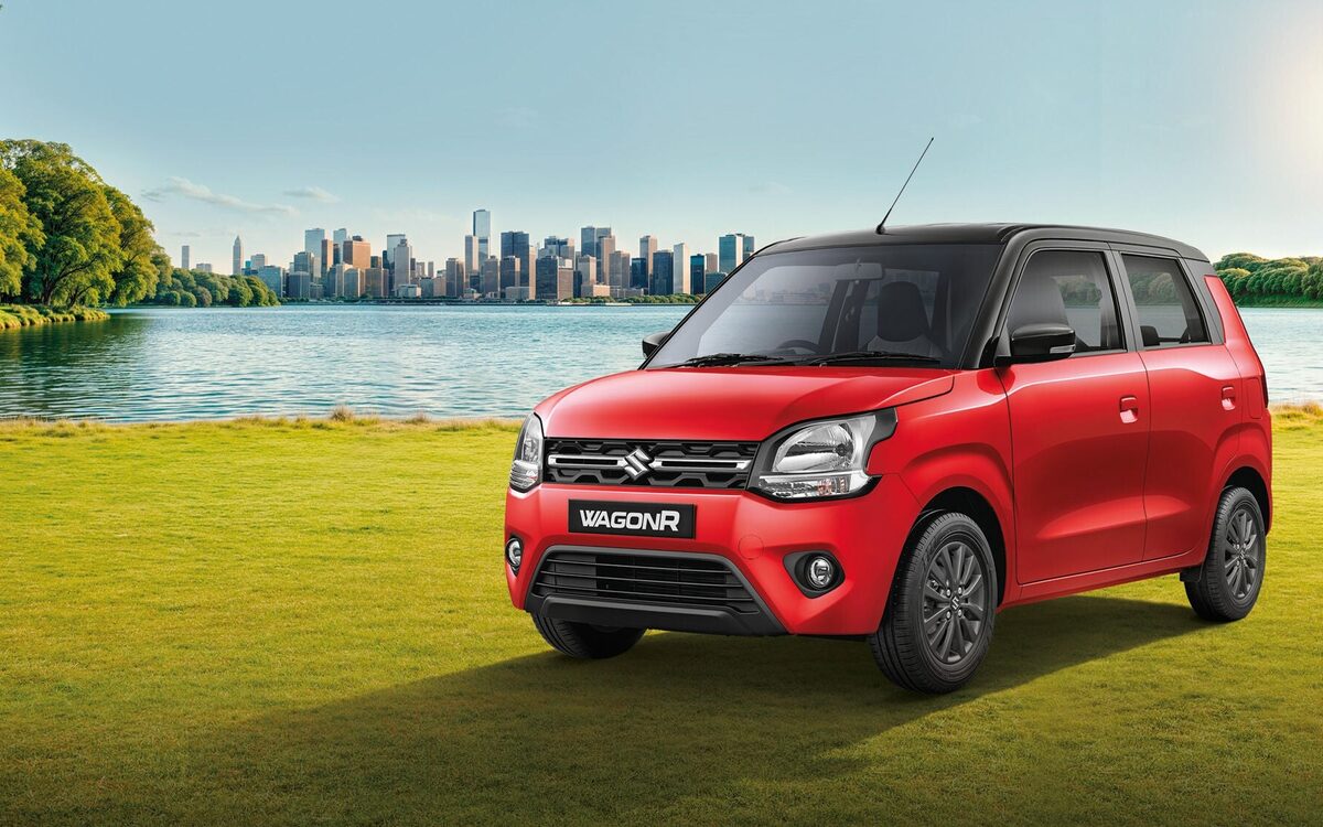Maruti Suzuki Wagon R in red with black roof, front three-quarter view parked on grass beside a lake with a city skyline behind