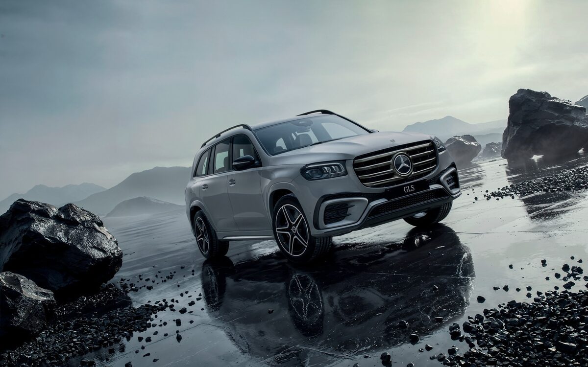 Mercedes-Benz GLS in silver parked on a wet rocky shore under a cloudy sky, front three-quarter view