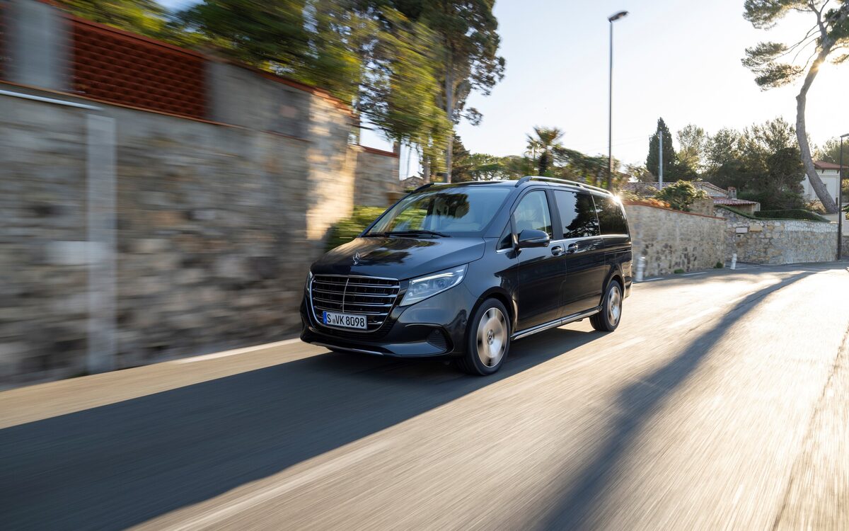 Black Mercedes-Benz V-Class driving along a sunny coastal road, front three-quarter view with motion blur on the road