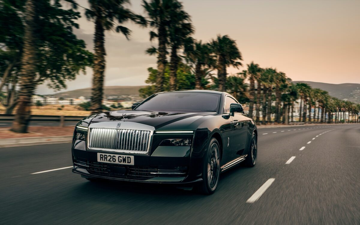 Rolls-Royce Spectre in dark green driving on a palm-lined coastal road, front three-quarter view