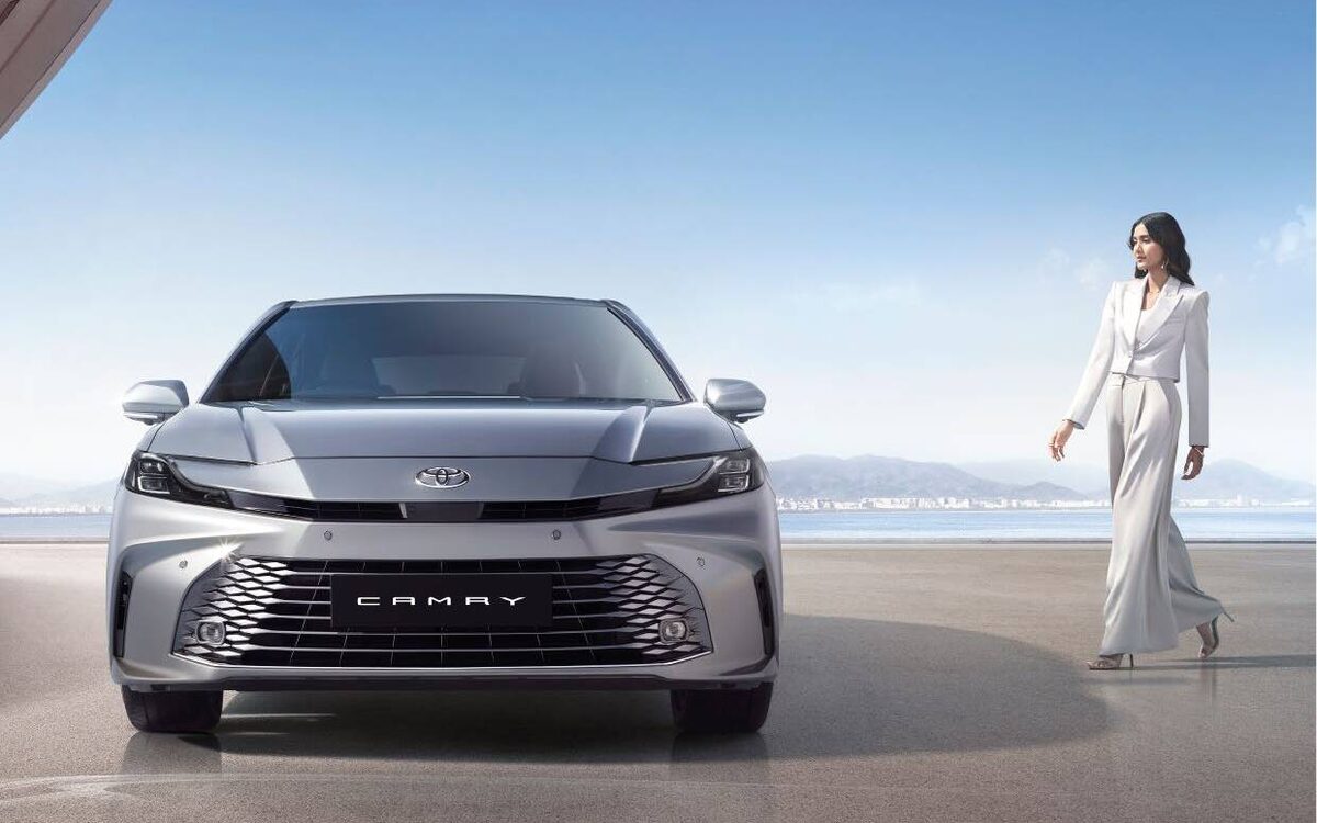 Grey Toyota Camry hybrid seen head-on with a woman in a white suit walking beside it