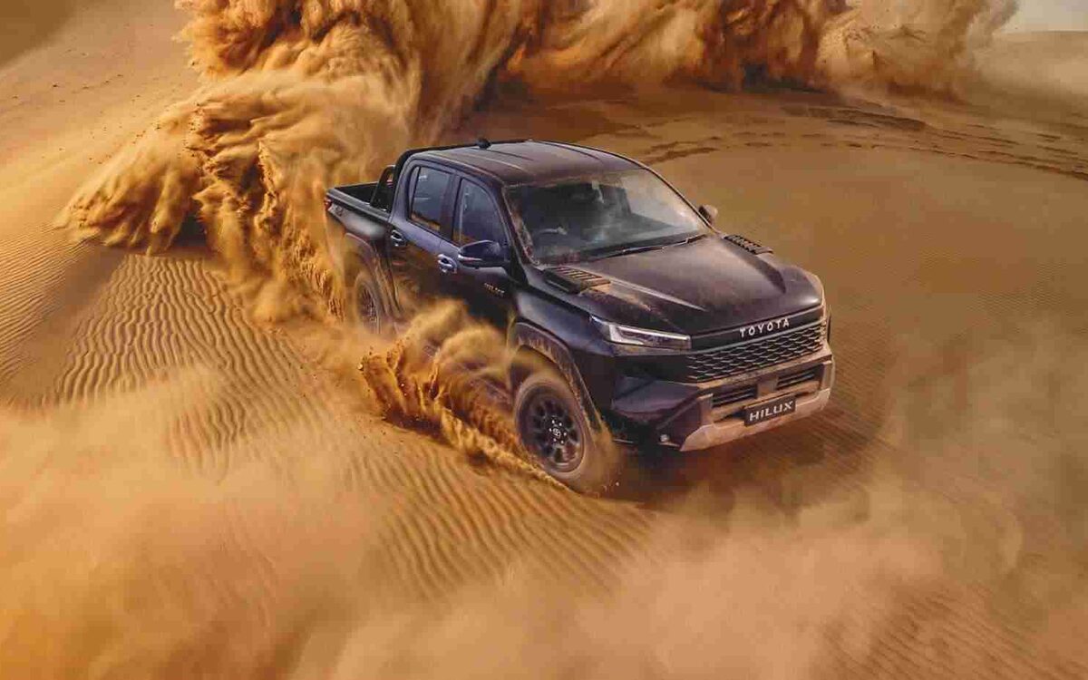 Dark-coloured Toyota Hilux driving through a sand dune kicking up a cloud of sand