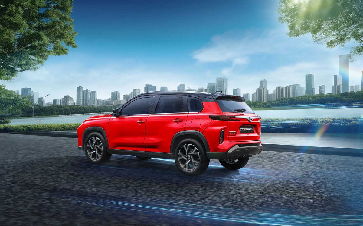 Toyota Urban Cruiser Hyryder in red, rear three-quarter view on a road beside a lake and city skyline