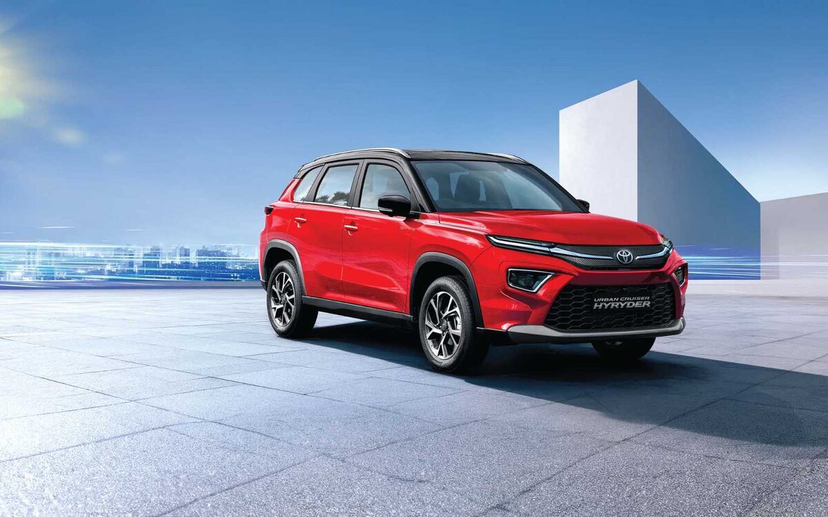 Toyota Urban Cruiser Hyryder in red with black roof, front three-quarter view on a paved area in bright sun