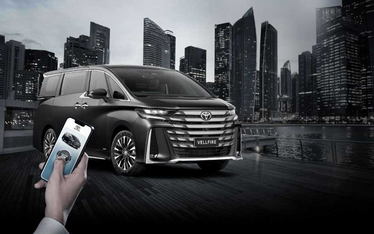 Toyota Vellfire in dark grey on a waterfront at night, with a hand holding a phone showing the car